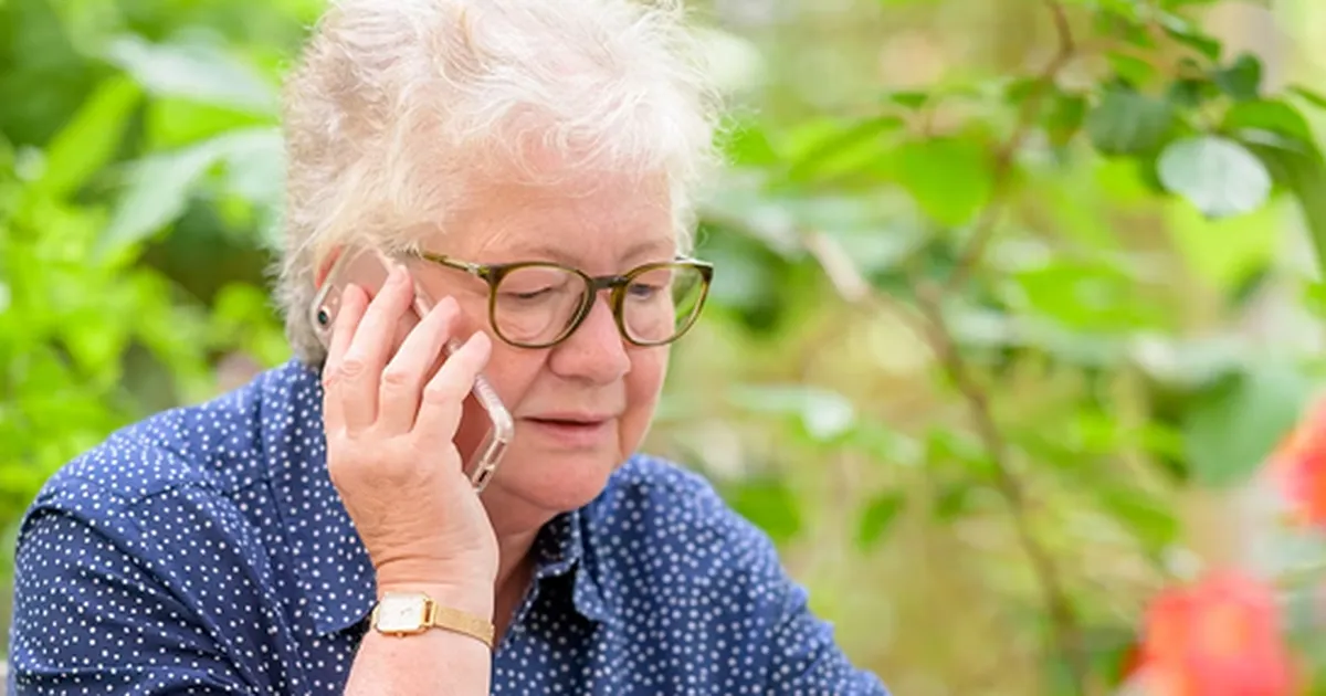 Une femme aînée aux cheveux blancs parle au téléphone devant son ordinateur, dans un jardin